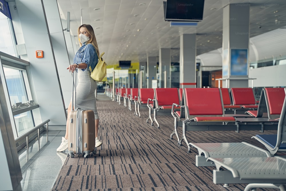 Cute long haired blonde putting her hand on the luggage and looking straight at camera, waiting for flight