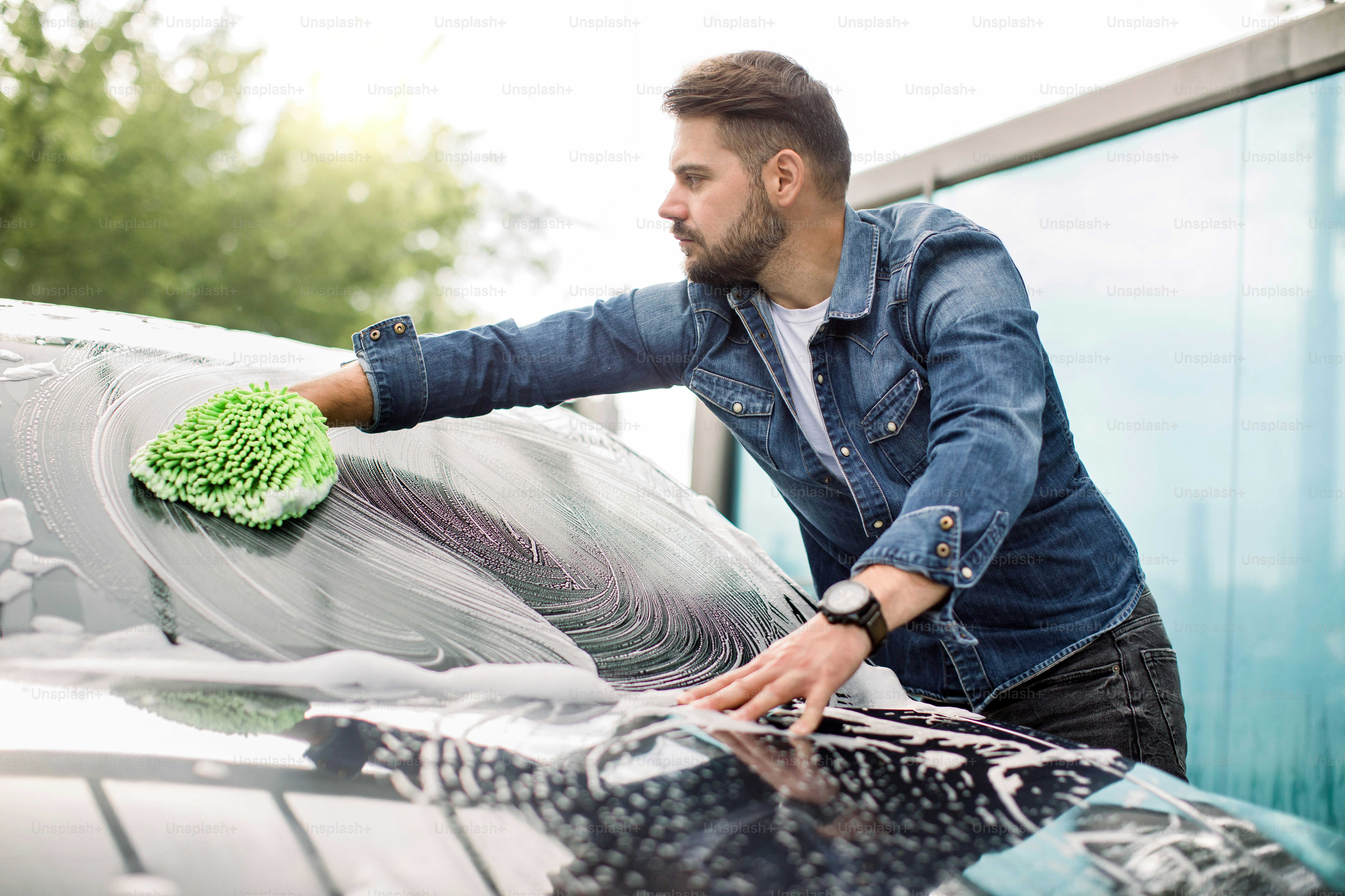 Horizontal view of car cleaning at self wash service outdoors. Handsome Caucasian man in casual wear washing the car windshield with green mitten sponge and foam