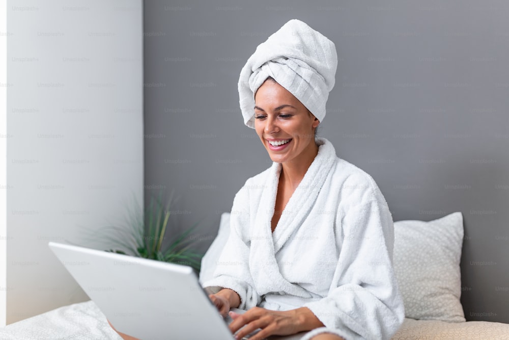 Young beautiful woman with towel on a bed in a hotel room, dressed in a white terry bathrobe, checking her email on her laptop computer, a slight smile on her face