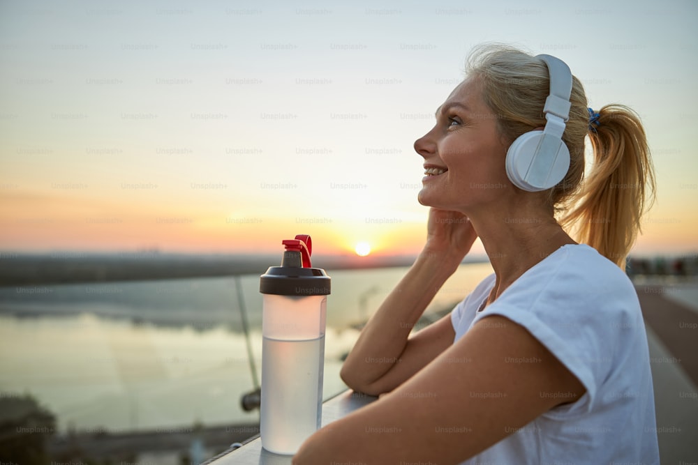 Vista lateral de uma mulher moderna de meia-idade sorridente satisfeita em fones de ouvido desfrutando de suas músicas favoritas
