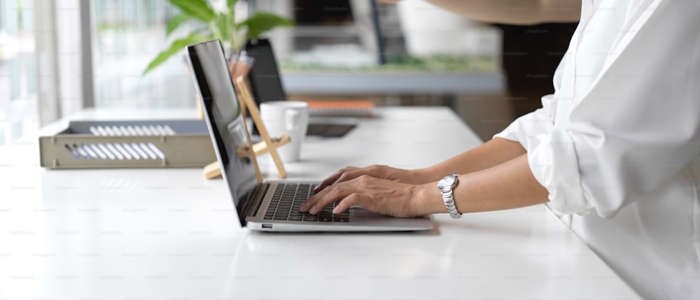 Vista lateral de manos femeninas escribiendo en el teclado de la computadora portátil en la mesa de trabajo con suministros de oficina en una sala de oficina moderna