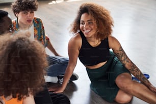 Happy young lady sitting on floor in dance studio while spending time with her friends