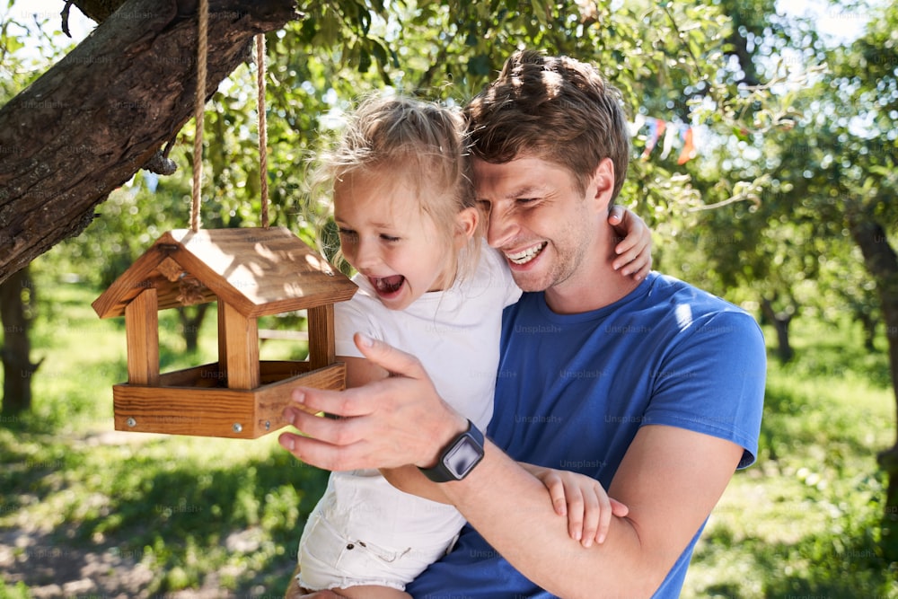 Joyful young man holding adorable little girl in his arms while looking at wooden birdhouse and laughing