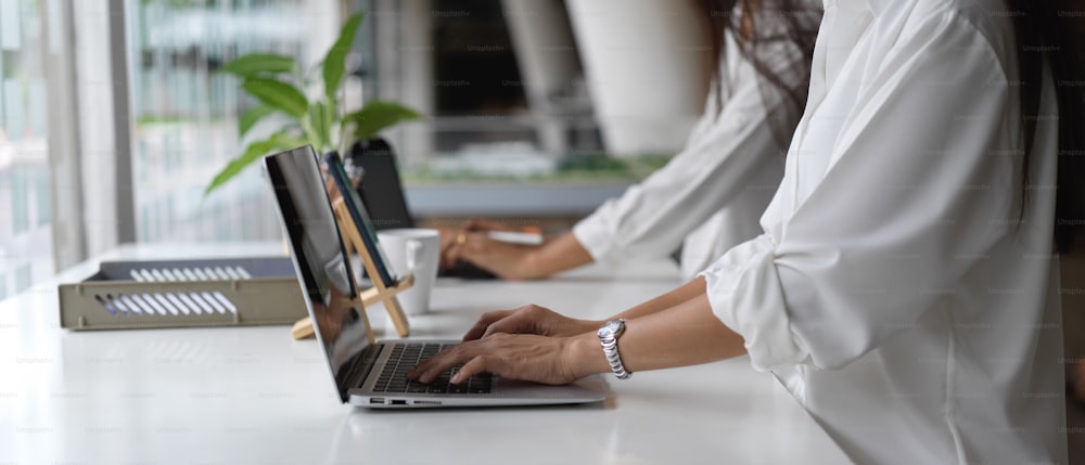 Side view of two female office workers working with laptop on office desk with office paper filing trays in office room