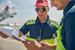 Supervisore concentrato della manutenzione degli aeromobili in abbigliamento da lavoro che fissa un documento nella mano del suo collega maschio