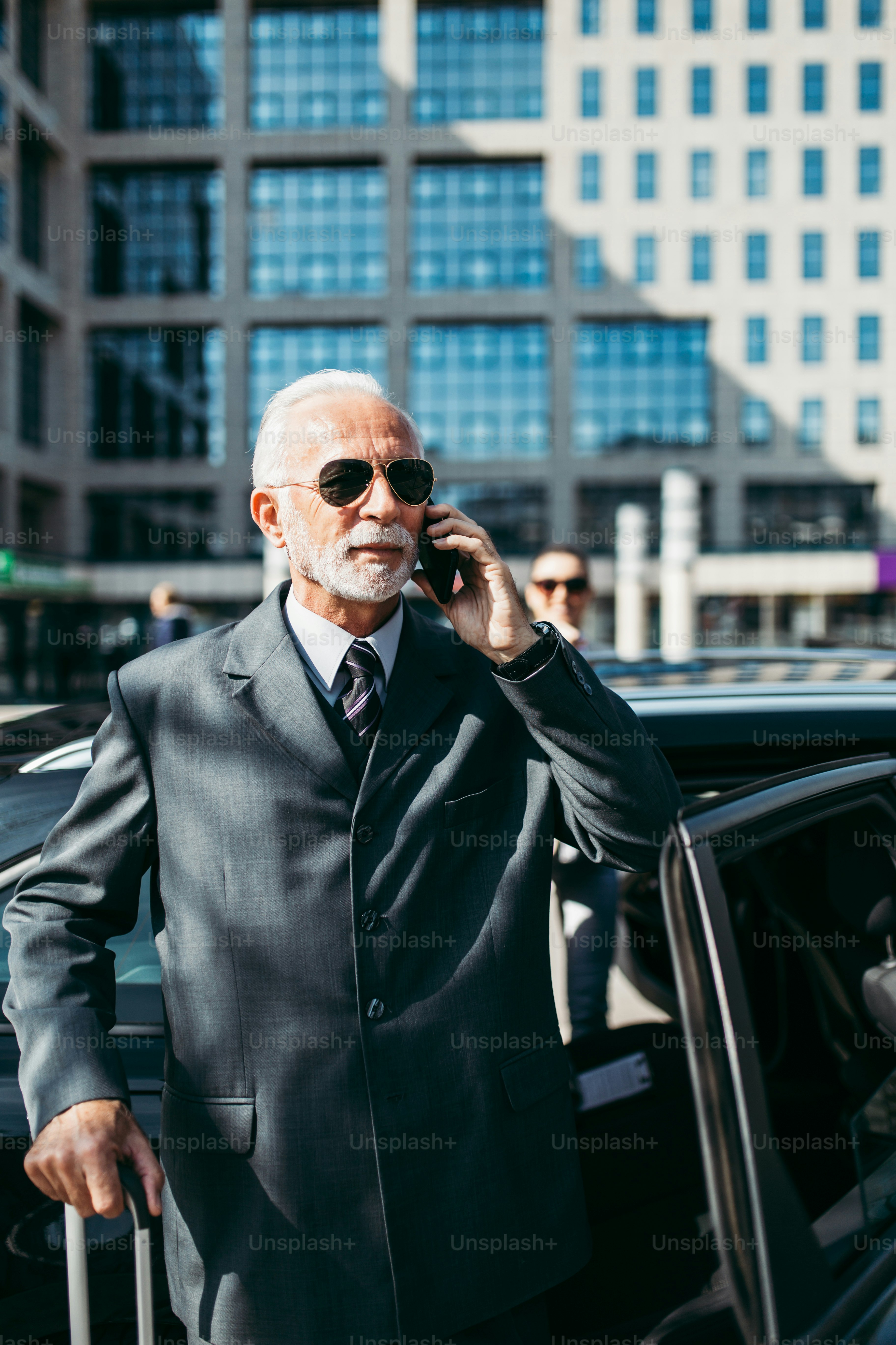 Good looking senior business man and his young woman colleague or coworker standing in front of luxury car. They talking, smiling and using laptop and smart phones. Transportation in corporate business concept.