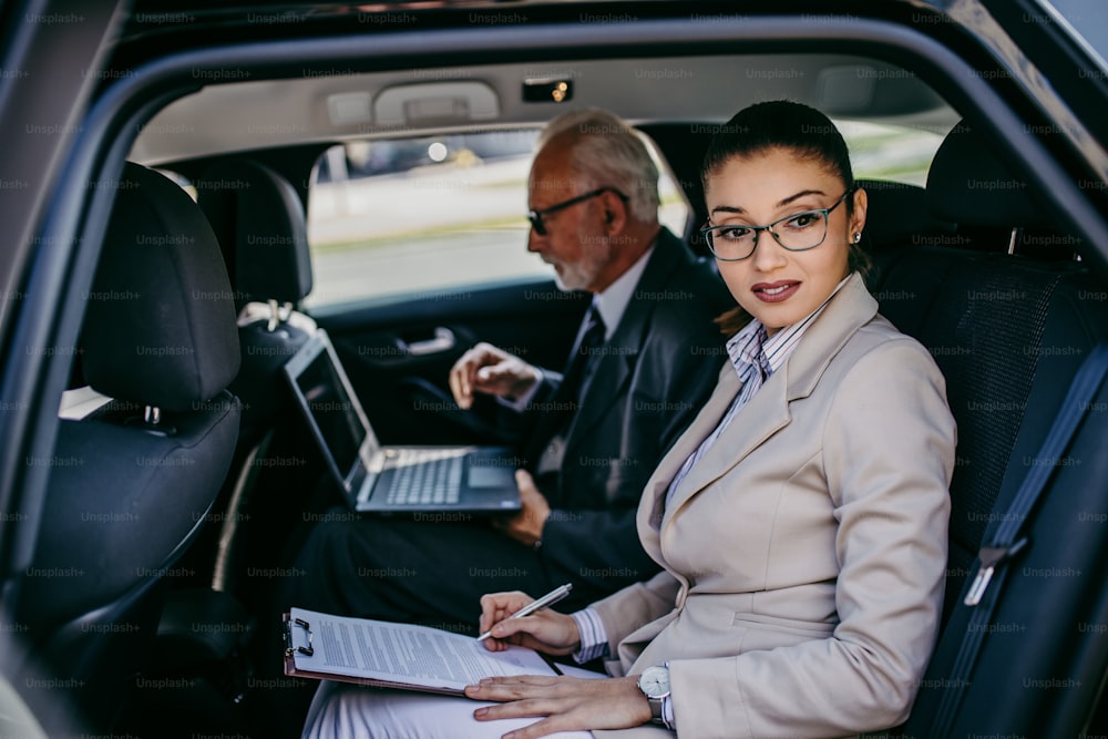 잘 생긴 고위 사업가와 그의 젊은 여성 동료 또는 동료가 고급 자동차의 뒷좌석에 앉아 있습니다. 그들은 이야기하고, 웃고, 노트북과 스마트 폰을 사용합니다. 기업 비즈니스 개념의 교통.