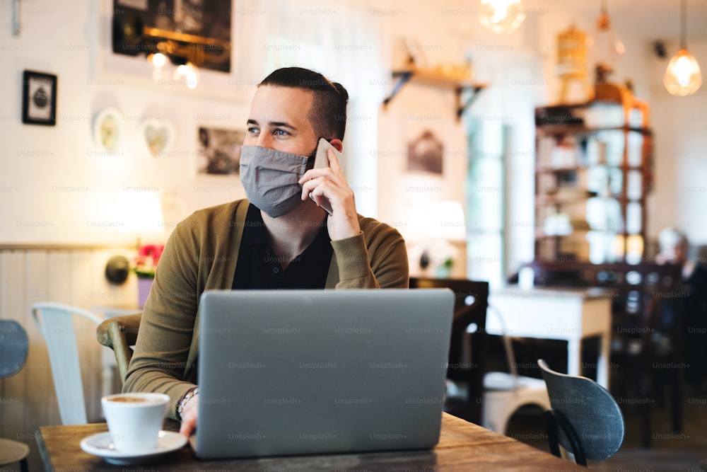 Portrait of young man with face mask and laptop indoors in cafe, using smartphone.