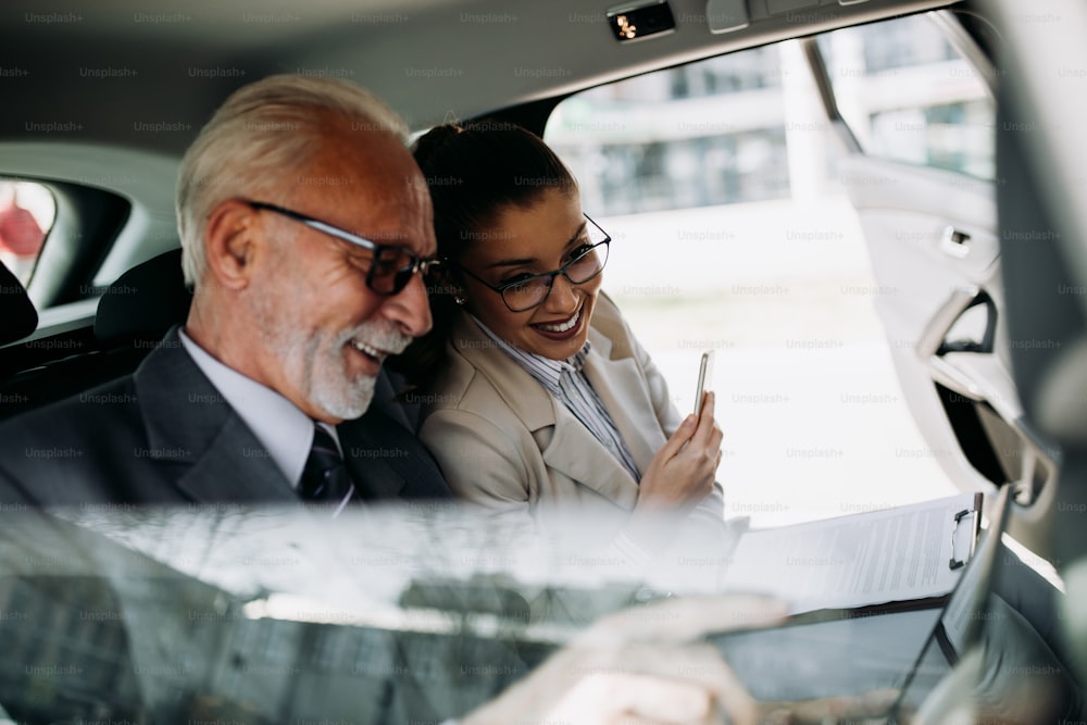 Good looking senior business man and his young woman colleague or coworker sitting on backseat in luxury car. They talking, smiling and using laptop and smart phones. Transportation in corporate business concept.