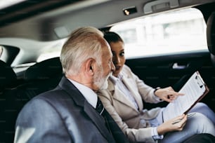 Good looking senior business man and his young woman colleague or coworker sitting on backseat in luxury car. They talking, smiling and using laptop and smart phones. Transportation in corporate business concept.