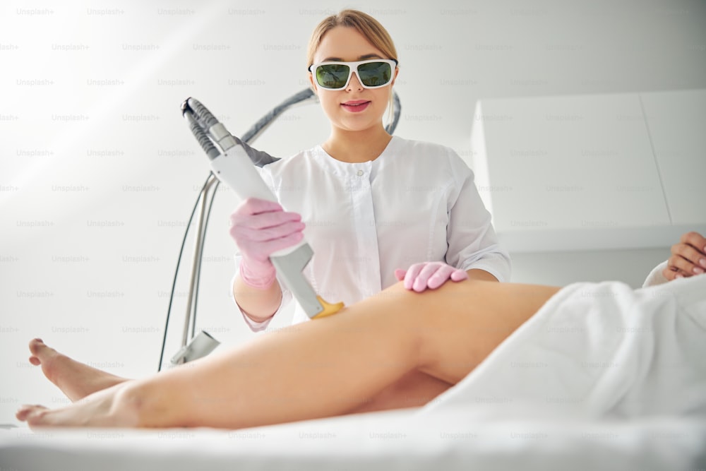 Concentrated professional cosmetologist in safety goggles performing a laser hair removal on a female patient leg