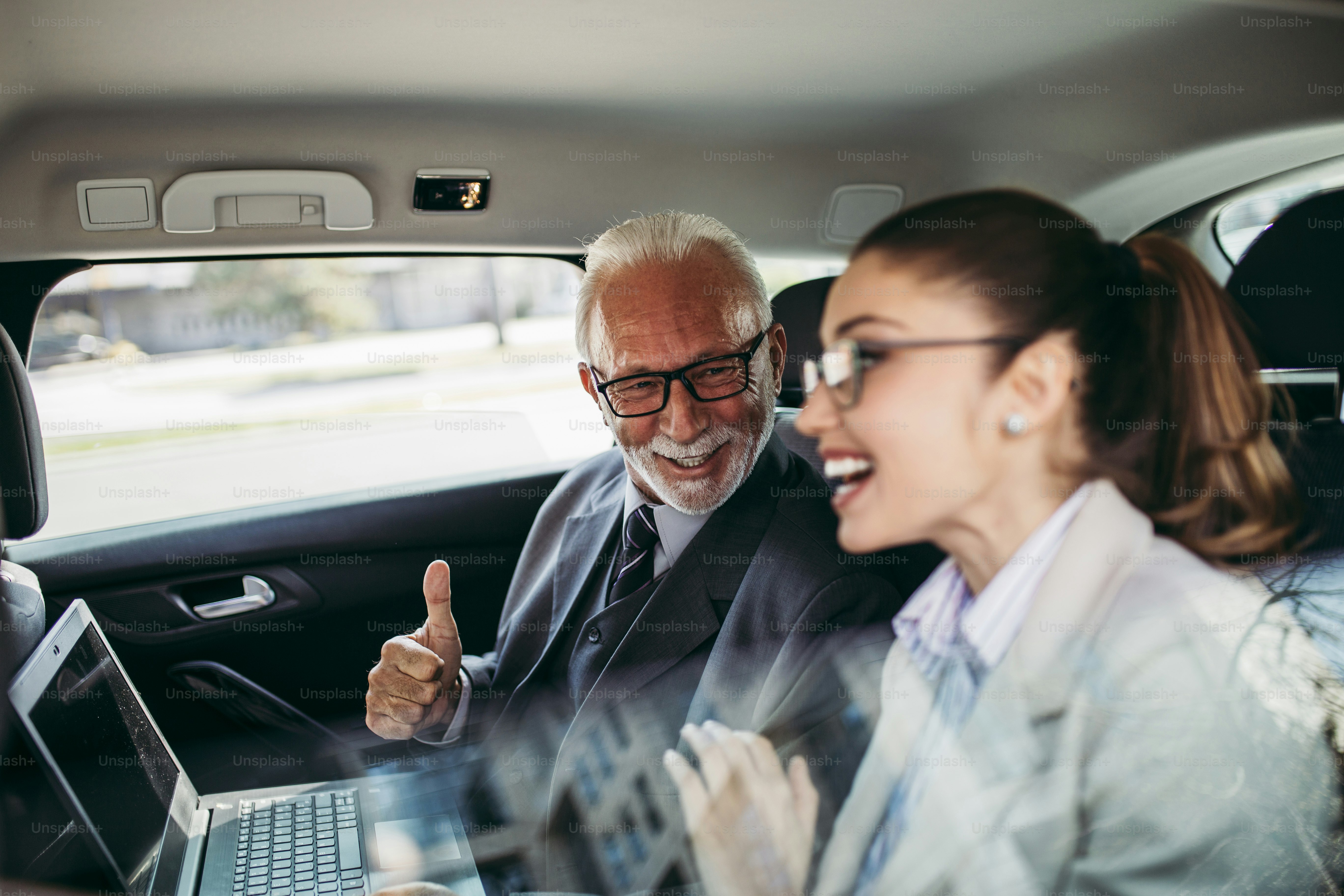 Good looking senior business man and his young woman colleague or coworker sitting on backseat in luxury car. They talking, smiling and using laptop and smart phones. Transportation in corporate business concept.