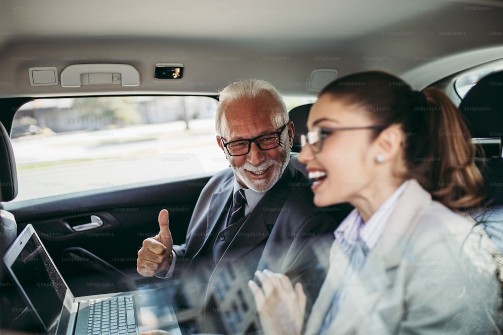 高級車の後部座席に座っている見栄えの良い年配のビジネスマンと彼の若い女性の同僚または同僚。彼らは話し、笑顔で、ラップトップやスマートフォンを使用しています。企業事業のコンセプトにおける輸送。