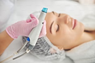 Female dermatologist holding a beauty device in her hand over her young patient with closed eyes