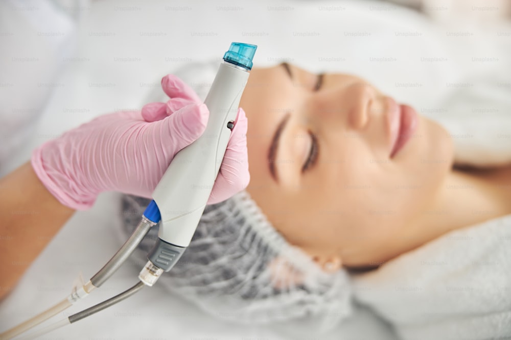 Dermatologista mulher segurando um dispositivo de beleza em sua mão sobre sua jovem paciente com os olhos fechados