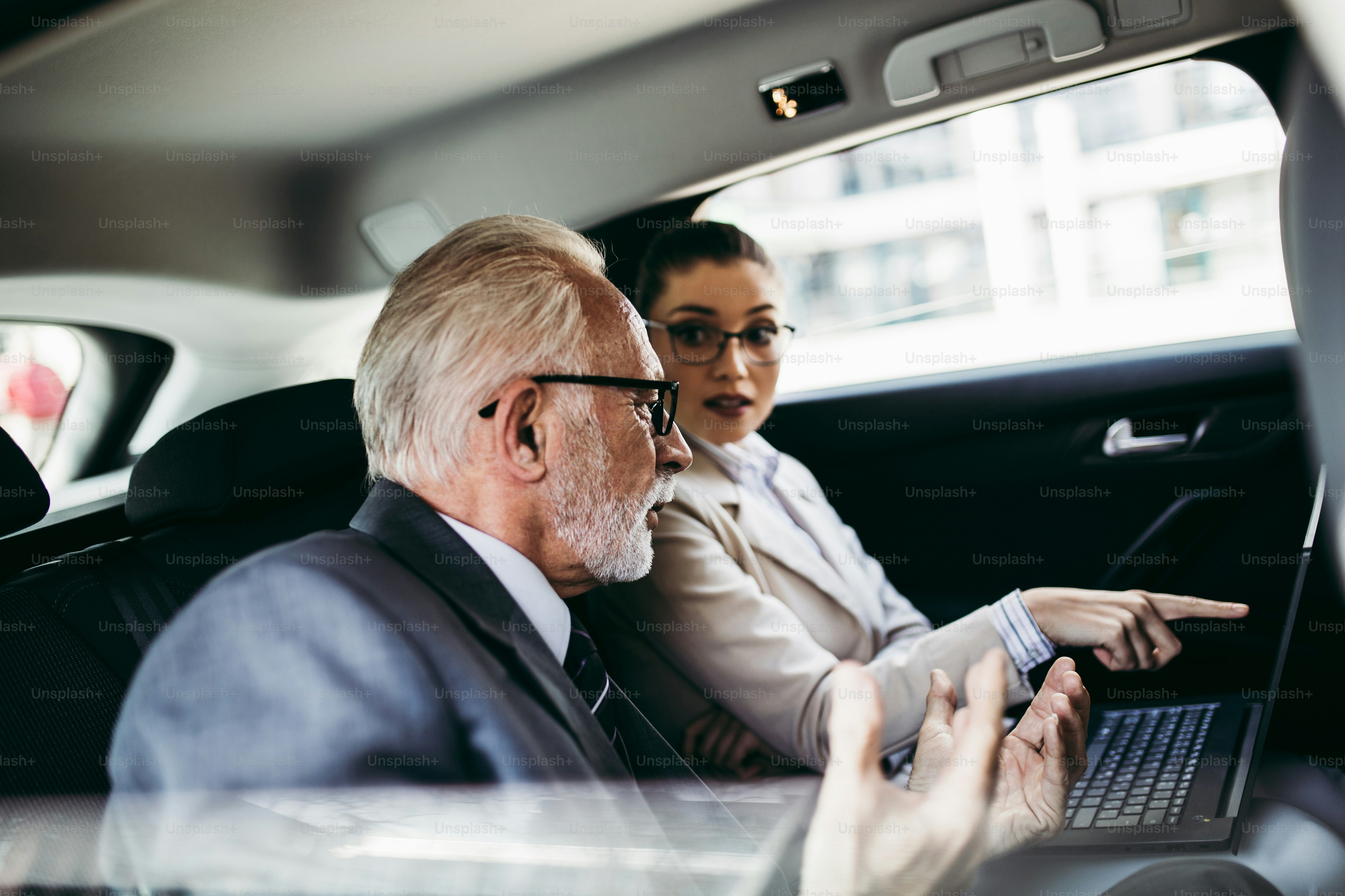 Good looking senior business man and his young woman colleague or coworker sitting on backseat in luxury car. They talking, smiling and using laptop and smart phones. Transportation in corporate business concept.