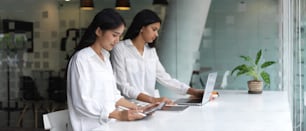 Side view of two female office workers working with digital devices on worktable in glass partition office room