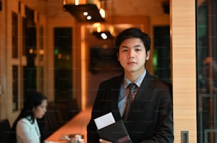 A businessman is holding a document file while standing over the meeting room as a background.