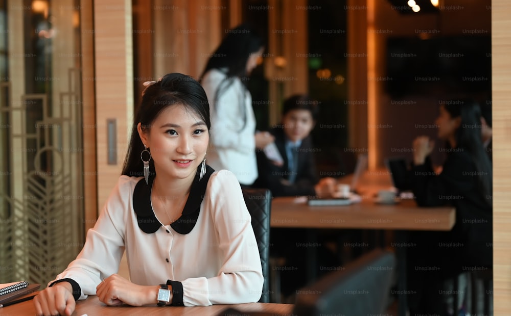 A businesswoman is sitting at the wooden working desk over a meeting room as a background.