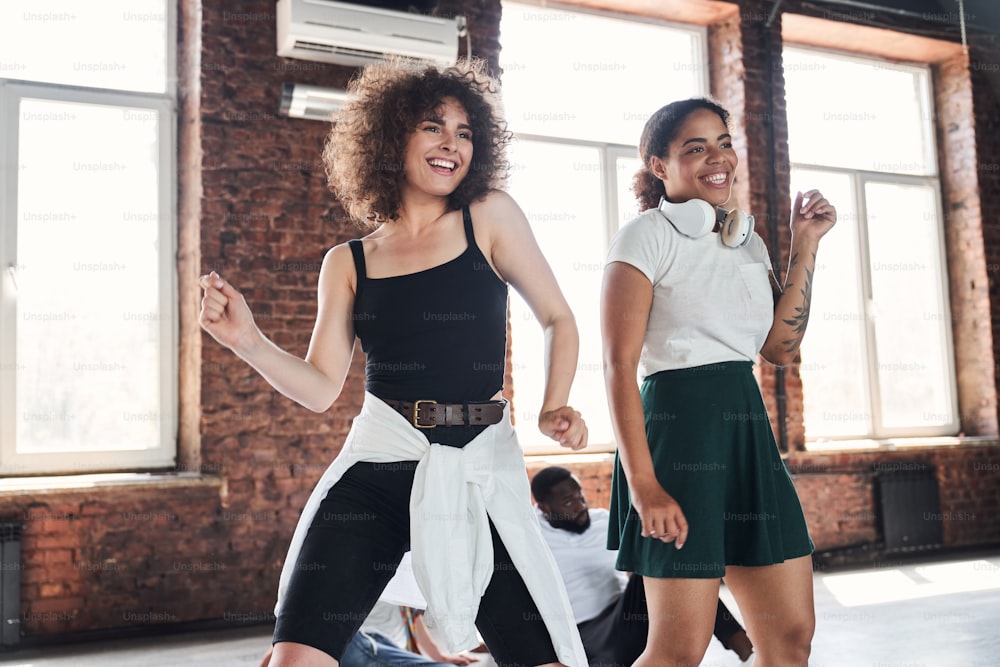 Two beautiful cheerful curly women are dancing freestyle together while training indoors with two guys