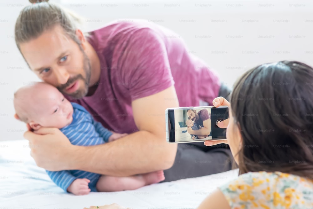 Caucasian woman smartphone taking a photo of her husband with sleeping newborn baby son on the bed.