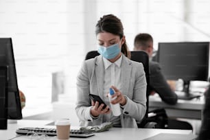 オフィスで電話を消毒する医療用マスクを着けたビジネスウーマン。