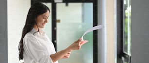 Eine schöne Frau prüft Papierkram, während sie über Bürowandglas steht.