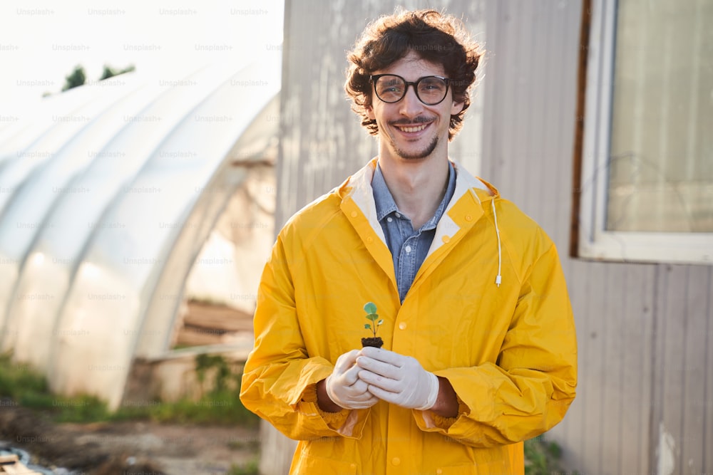 Waist up portrait of happy curly male in glasses standing on farm and holding small green sprout