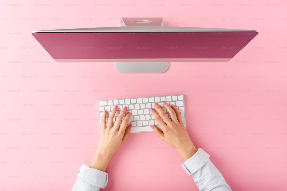 Office desktop concept. Woman’s hands working on computer on pink background. Top view