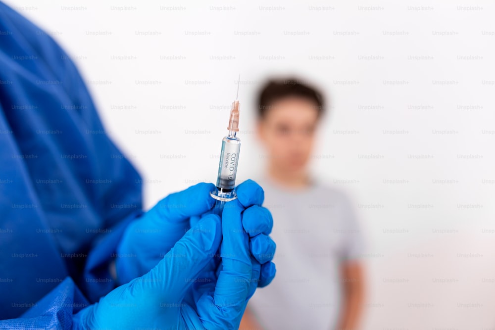 ワクチン接種。青い滅菌手袋をはめた専門医が注射器を持ち、医療用マスクを着けた小さな男の子に注射をしています。予防接種、医学、ヘルスケア。Covid19、コロナウイルスワクチン