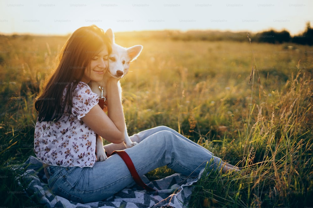 Happy woman hugging cute white puppy in summer meadow in sunset light. Atmospheric moment. Stylish girl relaxing with her adorable fluffy puppy on a picnic.  Summer trip with pet