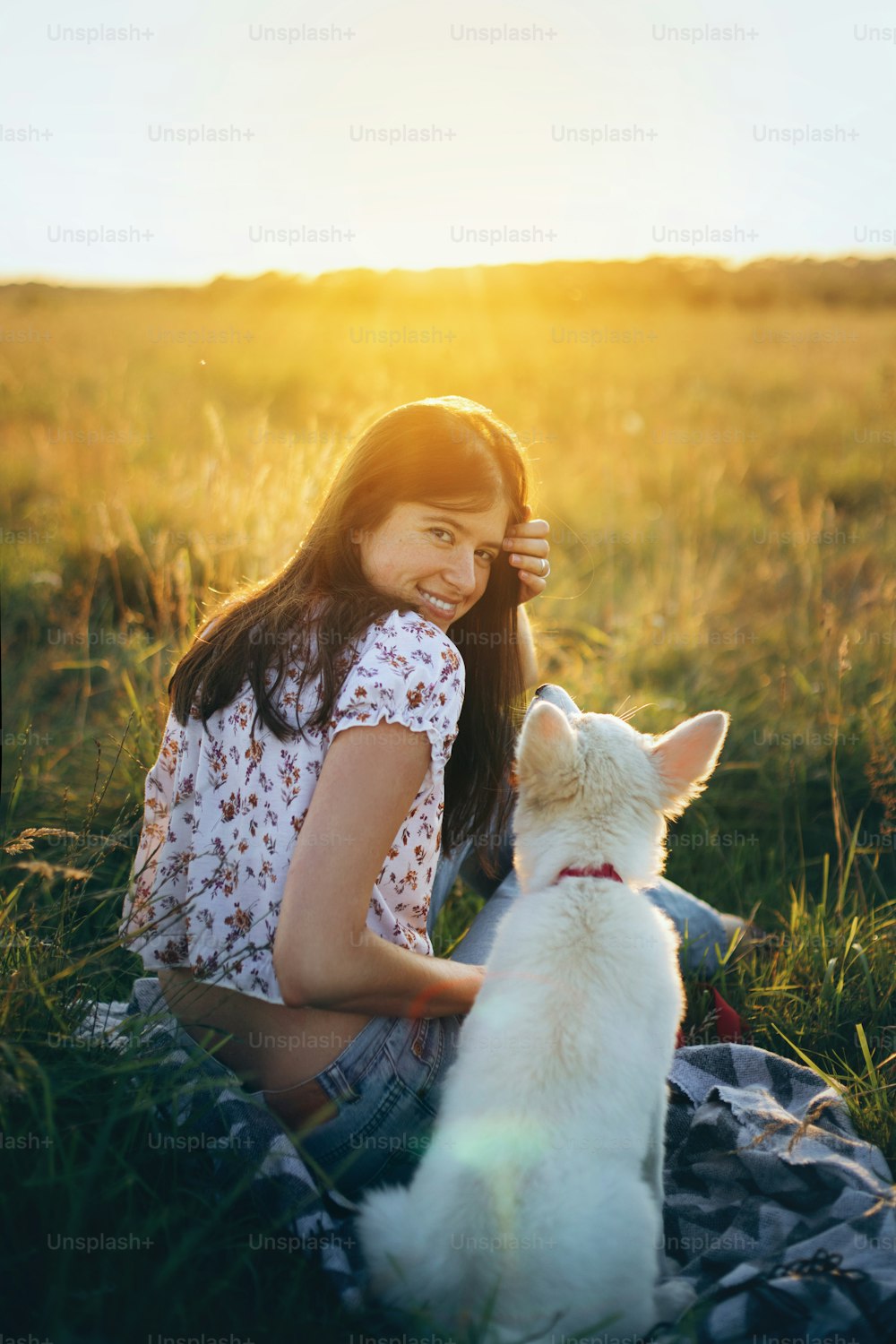 행복한 여자는 일몰 따뜻한 빛 아래 여름 초원에서 귀여운 하얀 강아지와 함께 앉아 있다. 피크닉에서 사랑스러운 푹신한 강아지와 함께 휴식을 취하는 세련된 소녀. 애완 동물과 함께하는 여름 여행