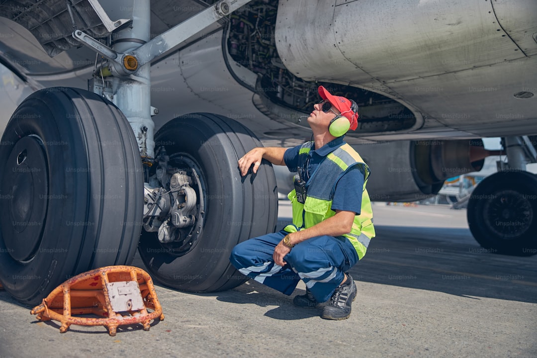 Serious aircraft maintenance mechanic in sunglasses and headphones sitting on his haunches under the plane