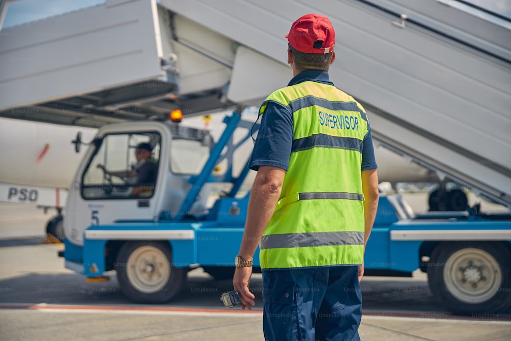 Back view of an airport worker supervising his colleague driving a truck with the passenger boarding stairs