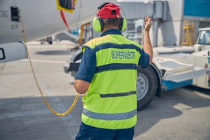 Back view of a skilled Caucasian aircraft maintenance supervisor in headset directing a pushback tractor operator