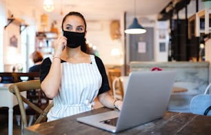 Portrait of young woman with apron, smartphone and laptop working in cafe.