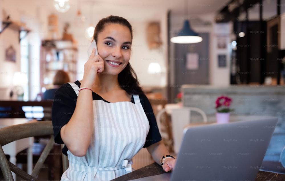 Retrato de una mujer joven con delantal, teléfono inteligente y computadora portátil trabajando en un café.