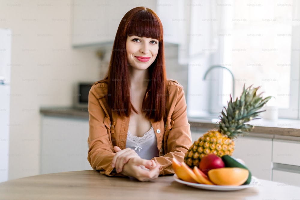 カジュアルなマスタードシャツを着たかなり若い美しい白人の赤毛の女性は、モダンなキッチンで新鮮な野菜や果物を持ってキッチンテーブルに座っています。健康的な食事とライフスタイルのコンセプト