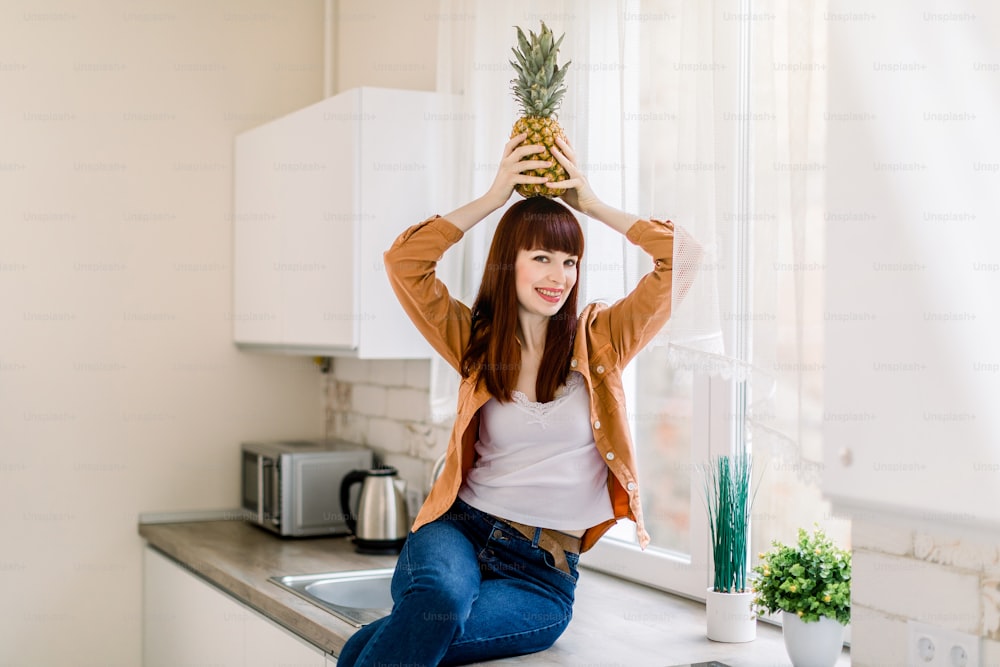 Foto de una hermosa joven en jeans y camisa informal, sosteniendo piña fresca en la cabeza y sonriendo a la cámara, posando en la cocina de casa sentada en la encimera. Comida sana, interior del hogar.