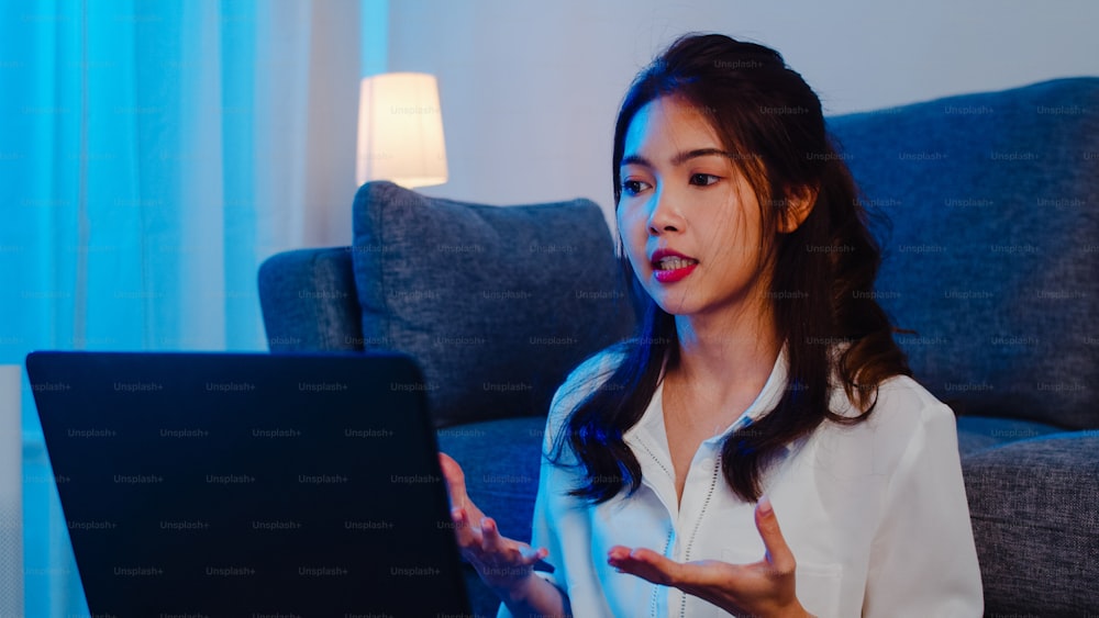 Asian businesswoman using laptop talk to colleagues about plan in video call while working from home at living room at night. Self-isolation, social distancing, quarantine for coronavirus prevention.