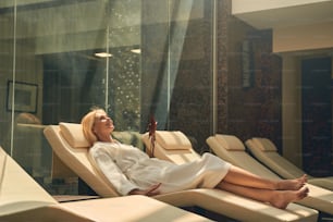 Full length portrait of charming lady in white soft bathrobe lying on deckchair in comfortable luxury spa hotel