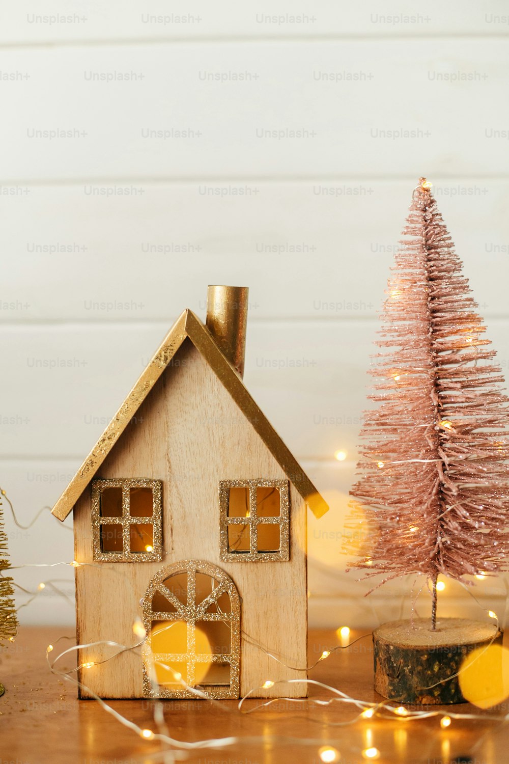 Stylish christmas house and glitter christmas tree in golden lights. Modern festive decor in scandinavian room. Miniature village with wooden house and tree toys. Merry Christmas