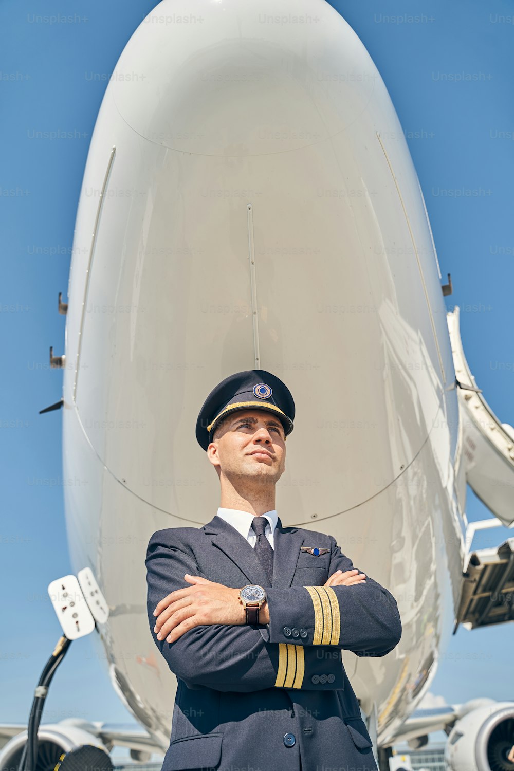 Angolo basso di un giovane aviatore pensieroso con le braccia conserte in piedi sotto la fusoliera dell'aereo