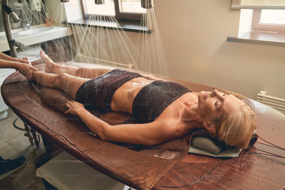 Full length portrait of charming woman lying on the massage desk while having shower rain on spa center