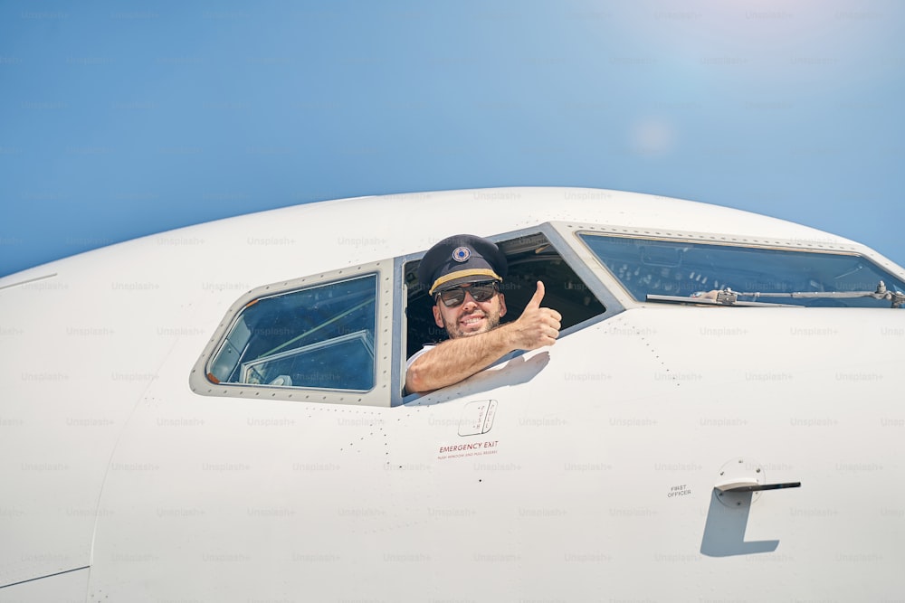 Contre-plongée d’un jeune homme souriant faisant un signe du pouce levé depuis la fenêtre de son cockpit