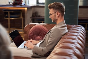 Serious entrepreneur wearing glasses sitting on a sofa in the hotel lobby and looking at the screen of a laptop on his laps