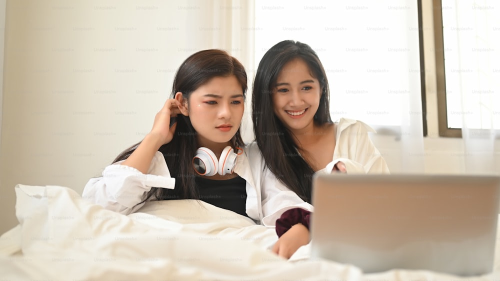 A Lesbian couple is watching a movie from a computer laptop while sitting together on the bed.