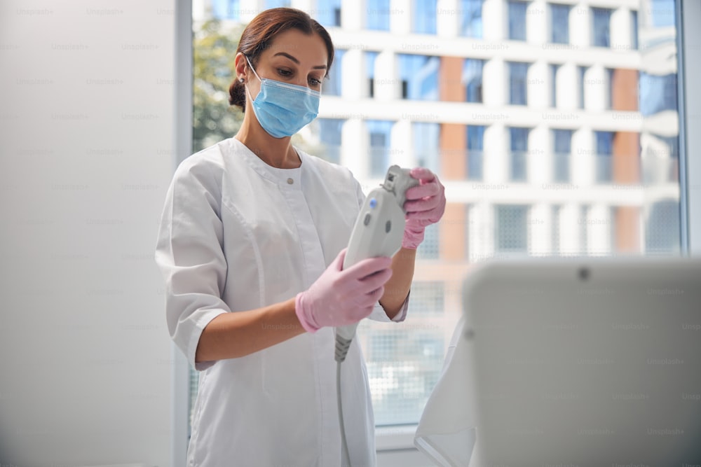 Zertifizierte Kosmetikerin in einer Gesichtsmaske, die eine Patrone in das Handstück eines Ultraschall-Gesichtsgeräts einführt