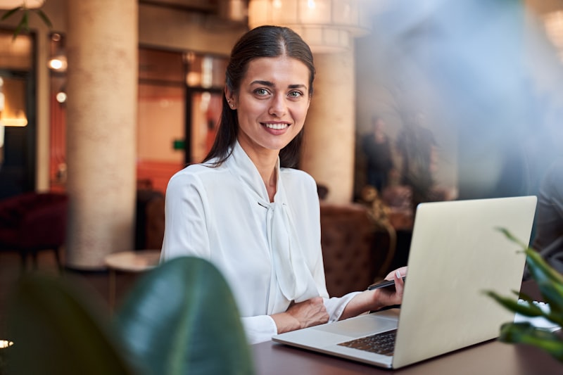 Confident attractive woman in a white blouse sitting at the bar counter with a smartphone in hand. Modern laptop in front of her