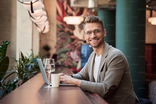 Positive handsome man sitting in a cafe with a modern laptop on a bar counter and smiling. Glass and paper cup by his side